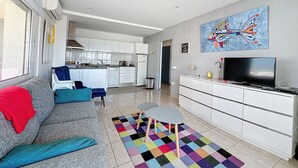 Ein modernes Wohnzimmer mit einer Couch, einem Couchtisch und einem Fernseher. Im Hintergrund befindet sich eine Küche mit weißen Schränken und einem Essbereich.
