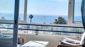 Ein Balkon mit Blick auf das Meer und Palmen, ausgestattet mit Korbstühlen und einem kleinen Tisch.