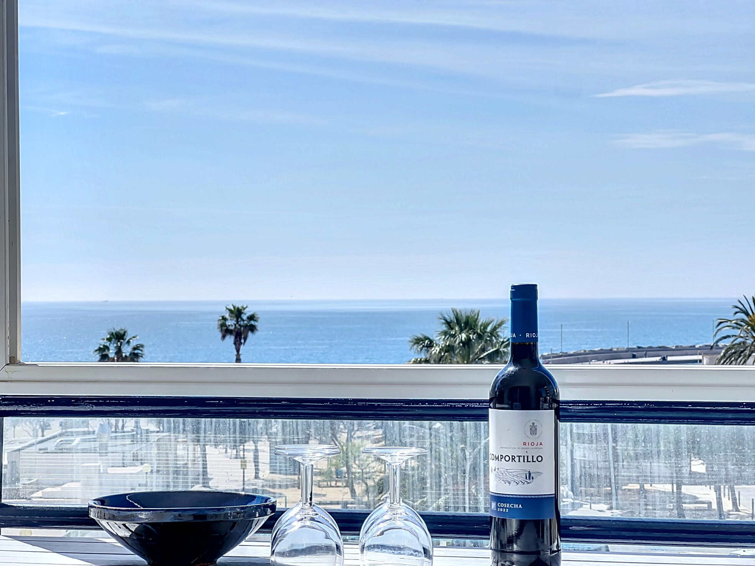 A bottle of Rioja wine, two wine glasses, and a black bowl on a table by a window overlooking the sea.