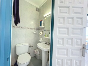 Casa de banho com sanita, lavatório e zona de duche. Há produtos de higiene pessoal no lavatório e uma toalha pendurada na parede.