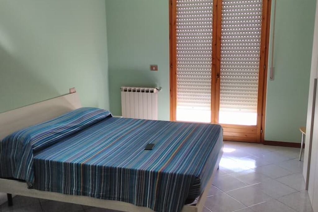 Een enkel bed met een gestreept beddegoed in een kamer met lichtgroene wanden, een radiator en een houten deur met zonwering.