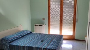Una cama individual con una colcha a rayas en una habitación con paredes verde claro, un radiador y una puerta de madera con persianas.