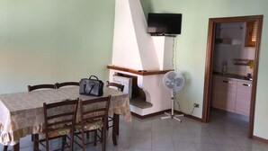 Un área de comedor con una mesa, sillas, un televisor montado en la pared, un ventilador y una cocina visible a través de una puerta abierta.