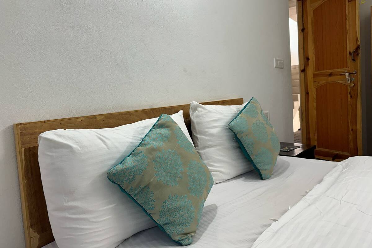 Ein Bett mit weißer Bettwäsche und zwei Kissen mit blaugrünem Muster, ein Holz-Kopfschild und eine Holztür im Hintergrund.