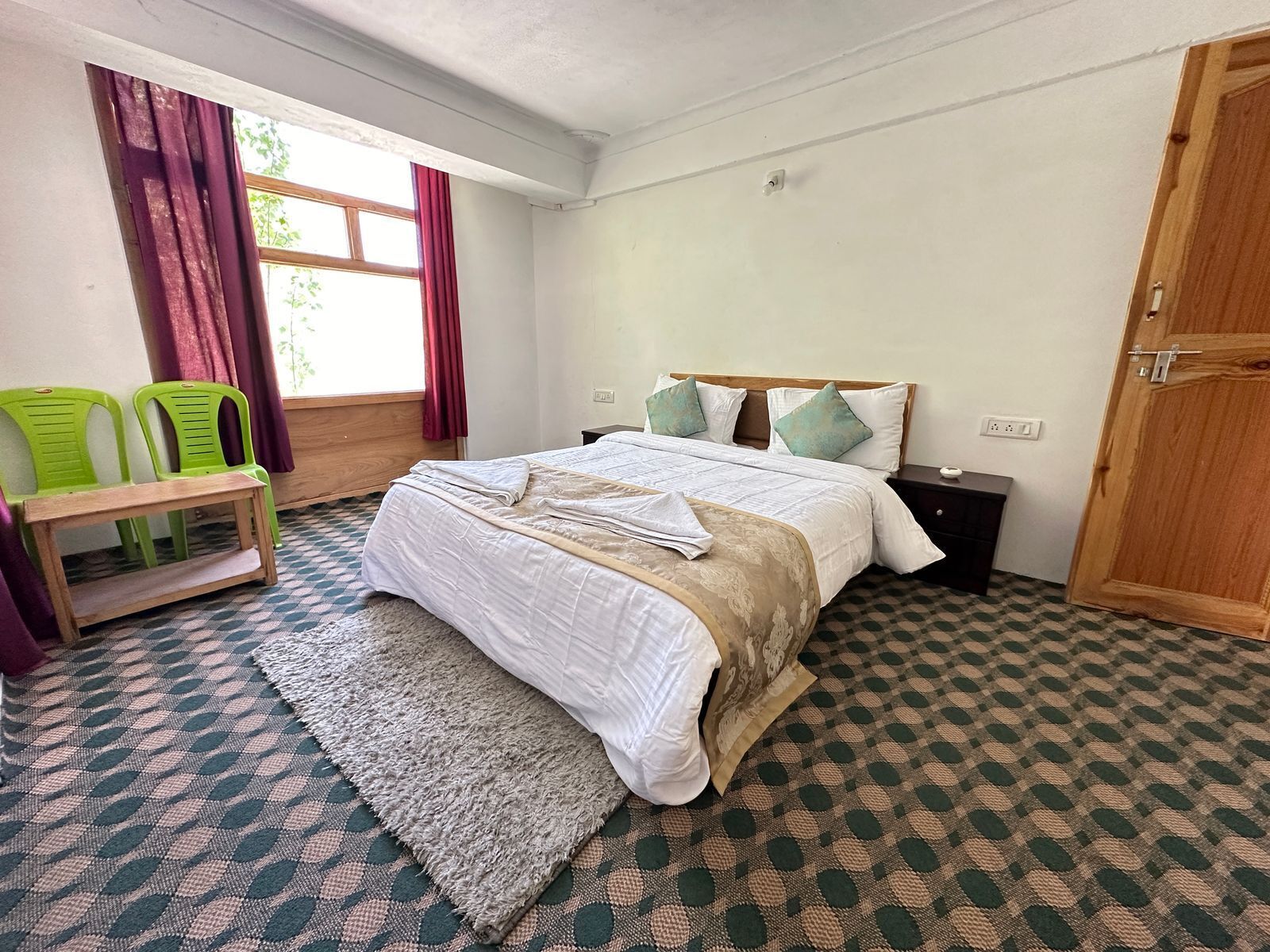 Un dormitorio con una cama, una mesita de noche, un escritorio, dos sillas, una ventana con cortinas y una puerta de madera.