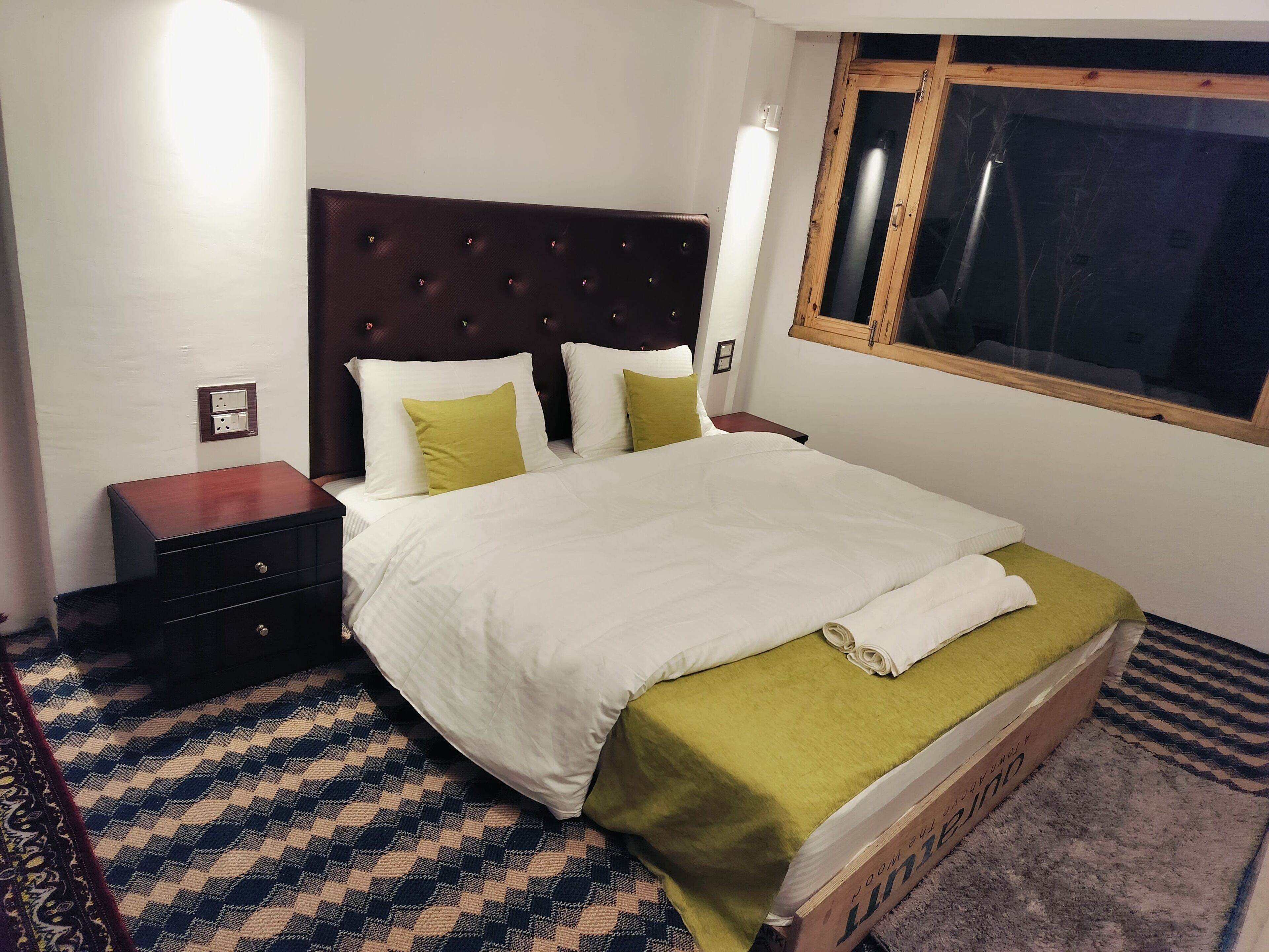 Uma cama bem arrumada com colcha branca e verde, cabeceira de madeira e uma pequena mesa de cabeceira com abajur.
