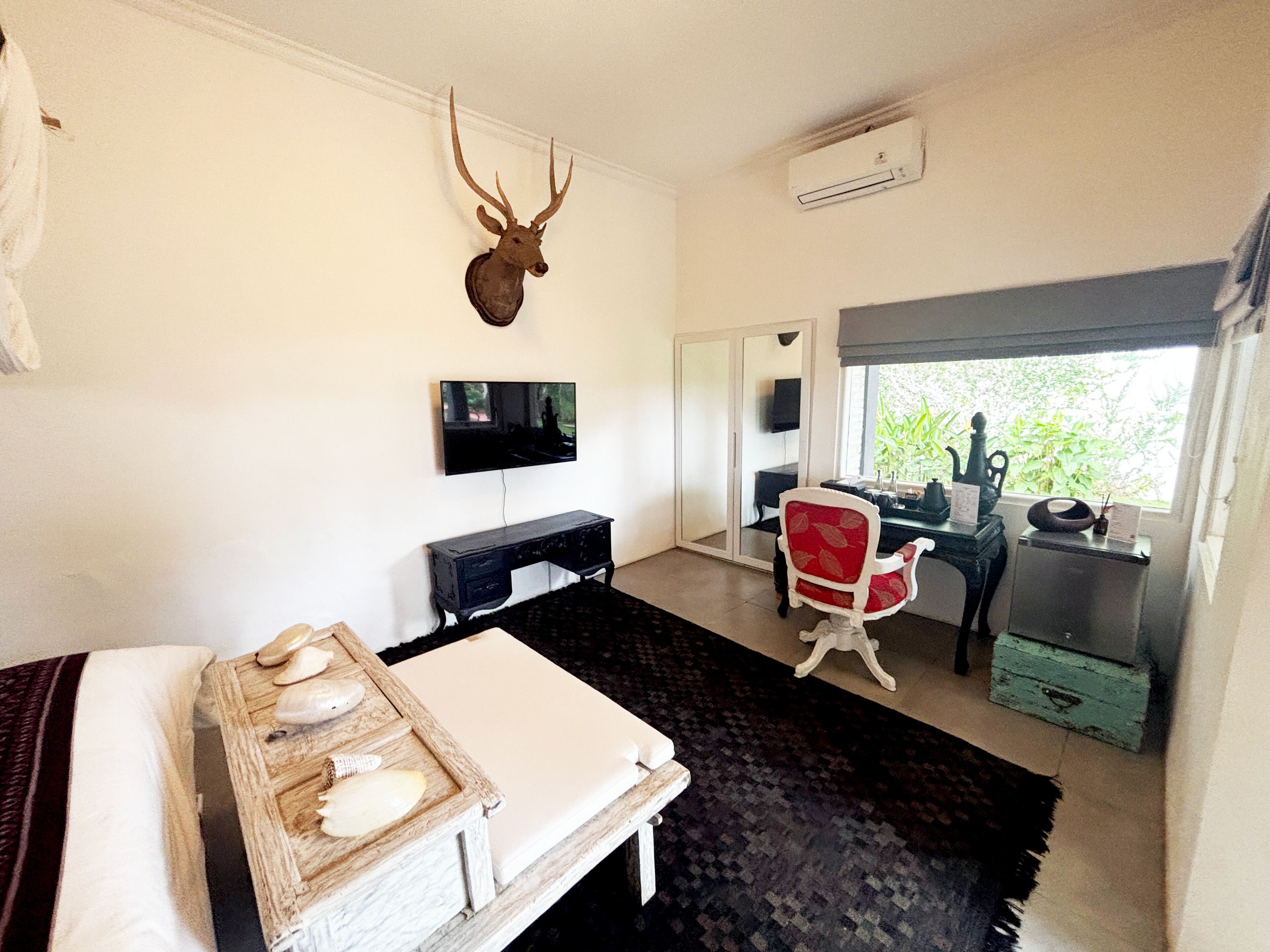 A room with a bed, a mounted deer head, a desk with a computer, a red chair, and a window with a view of greenery.