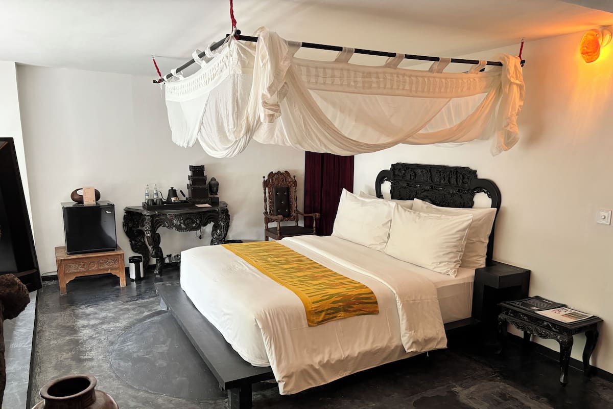 A bedroom with a canopy bed, a bedside table, a nightstand, a decorative chair, and a small table with items on it.