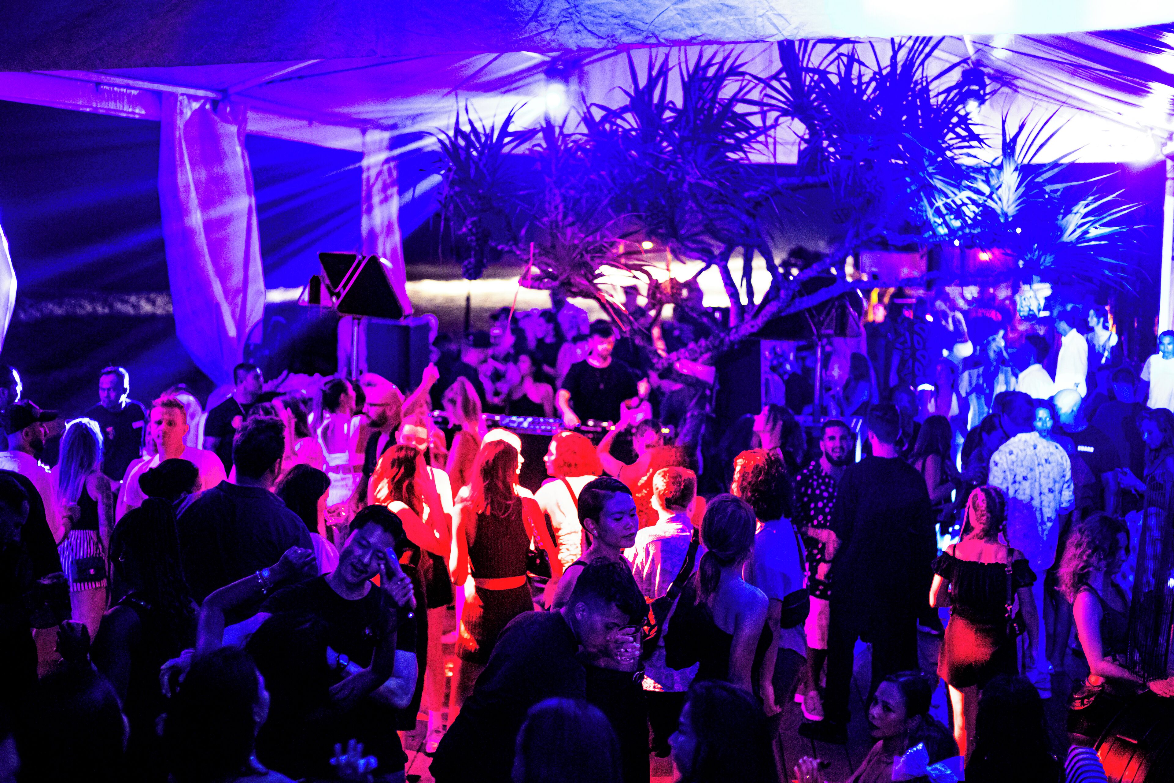 Un evento interior concurrido, con gente interactuando y un puesto de DJ al fondo.