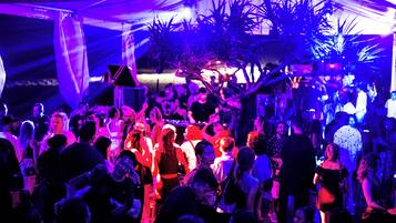 Un evento interior concurrido, con gente interactuando y un puesto de DJ al fondo.
