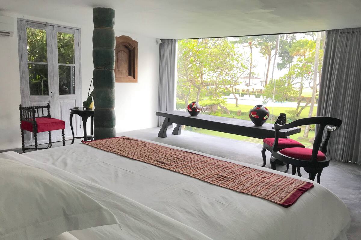 A bedroom with a large bed, a bench, two chairs, a window with curtains, and a view of greenery outside.