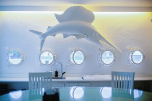 A room with a large table, four porthole windows, a white wall, and a ceiling-mounted fish sculpture. - Morabito Art Villa (Tibubeneng)