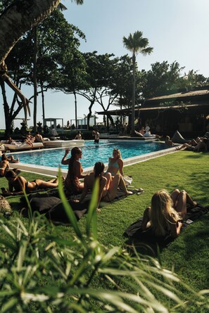 A poolside area with people relaxing on grass and lounge chairs, surrounded by trees and a clear sky. - Morabito Art Villa (Tibubeneng)