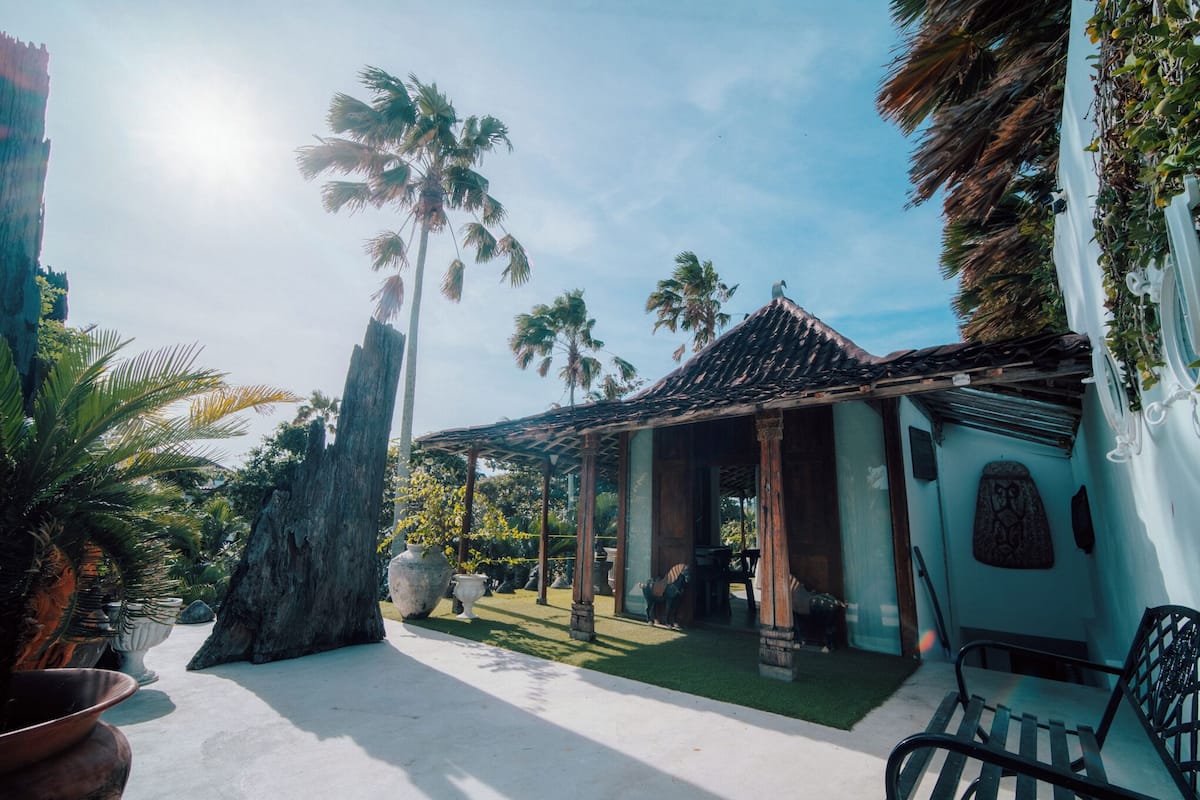 A tropical resort with a covered outdoor seating area, palm trees, and a large rock centerpiece.