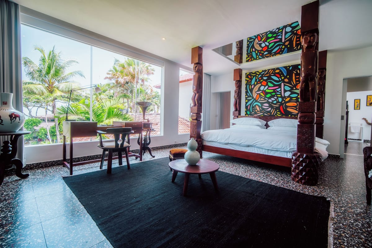 A hotel room with a large bed, a dining area with a table and chairs, a decorative vase, and a view of palm trees outside.