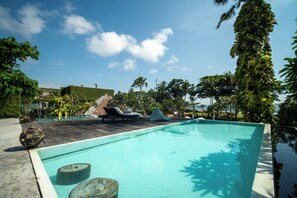 A swimming pool with lounge chairs and a wooden deck, surrounded by tropical vegetation. - Morabito Art Villa (Tibubeneng)