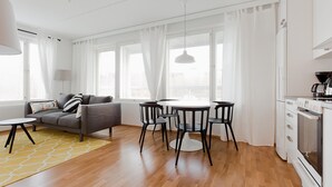 A modern living room with a grey sofa, a round dining table with black chairs, a yellow patterned rug, and a white kitchen with a stove. - Forenom Apts Turku Laukkavuori (Turku)