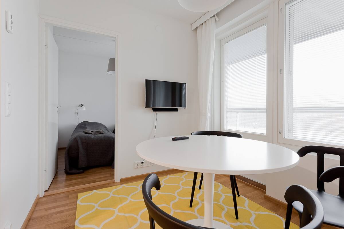 Ein Zimmer mit einem runden weißen Tisch, schwarzen Stühlen, einem flachbildfernseher an der Wand und einem Bett im Hintergrund.