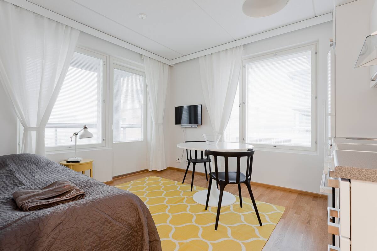 Ein Hotelzimmer mit einem Bett, einem kleinen Tisch mit Stühlen, einem Fernseher und großen Fenstern mit Vorhängen.