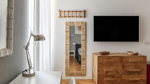A modern room with a wooden cabinet, a desk with a lamp, a flat-screen TV, and a mirror reflecting a room with a sofa and a chair. - Piazza Vittorio Luxury House (Roma)