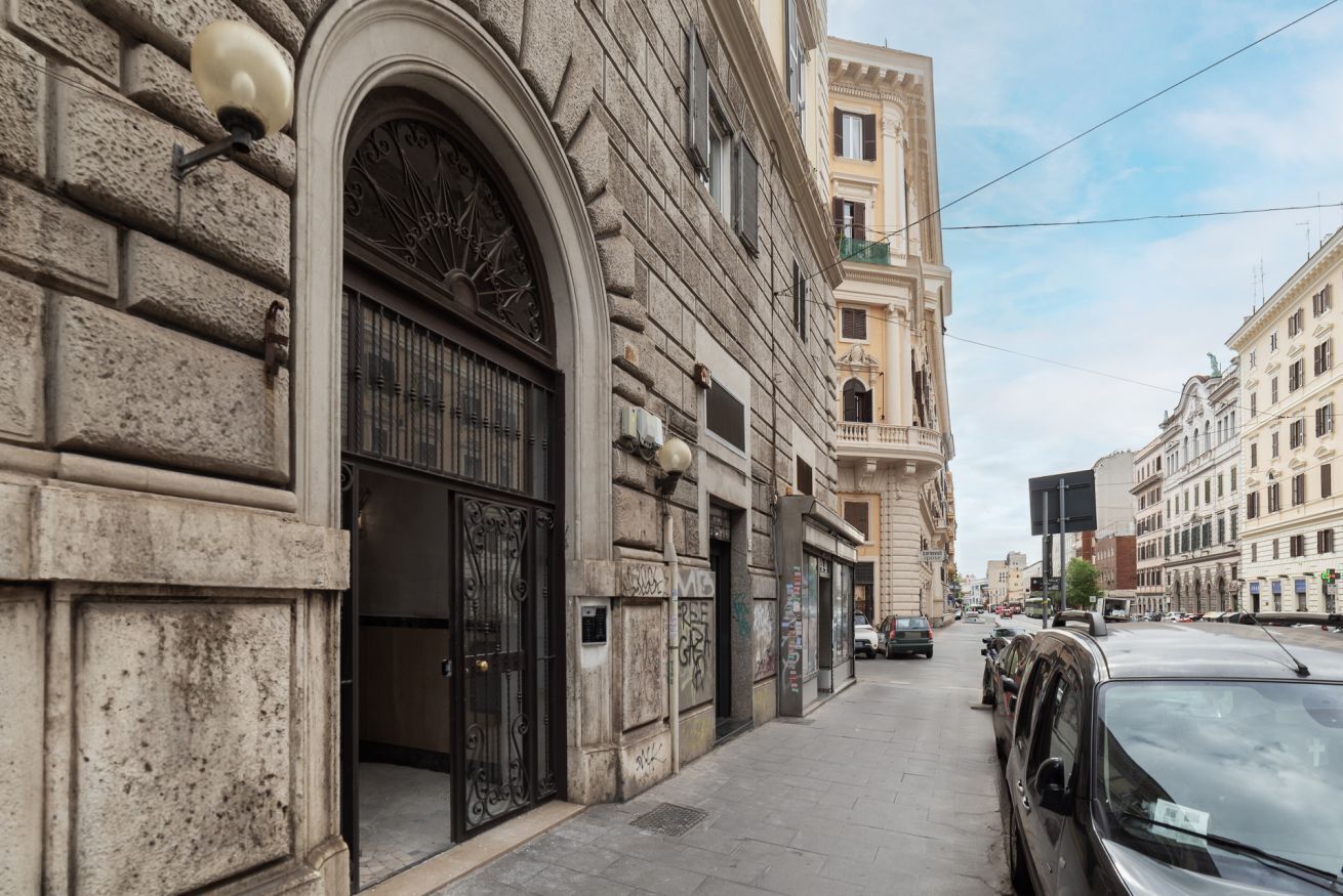 A street view with a building featuring a large arched window and a door, adjacent to parked cars and other buildings.