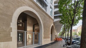 Un edificio con amplias ventanas de arco, una entrada vigilada y un paseo pavimentado con coches aparcados y árboles.