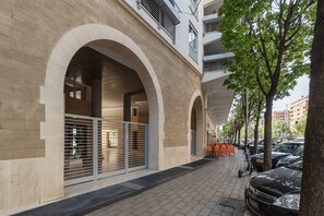 A building with large arched windows, a gated entrance, and a paved sidewalk with parked cars and trees.