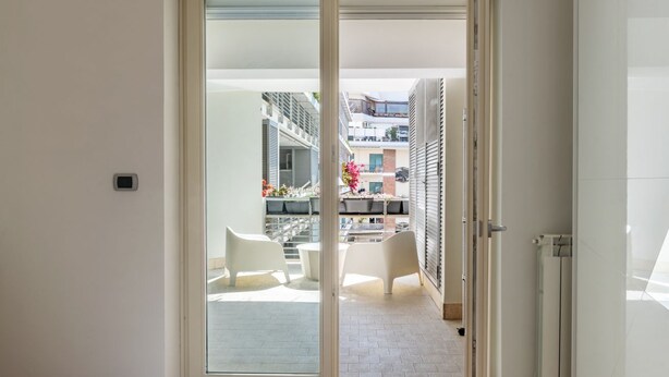 Habitación de hotel con puerta corrediza de vidrio que da a un balcón exterior con sillas blancas y vistas de edificios cercanos.