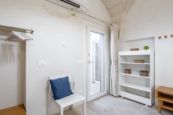 Una habitación con un banco blanco, una estantería con cestas, una puerta con panel de vidrio y un equipo de aire acondicionado.