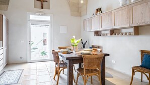 Een eethoek met een tafel voor vier personen, omringd door rieten stoelen, een houten kast erboven en een kruis aan de muur.