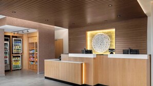 A modern reception desk with a wooden finish, a decorative wall piece, and a refrigerated display area with snacks. - Fairfield by Marriott Inn & Suites Kansas City at The Legends (Kansas City)