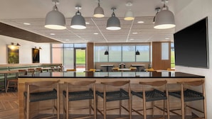 A modern bar area with a long counter, multiple bar stools, pendant lighting, and large windows offering views of the outdoors. - Fairfield by Marriott Inn & Suites Kansas City at The Legends (Kansas City)