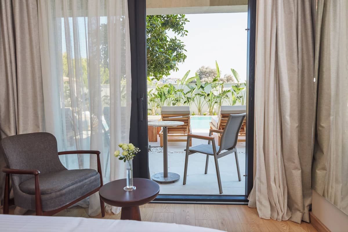 Une chambre d’hôtel avec un lit, une chaise, une petite table ornée d’un vase de fleurs, et une vue sur un espace extérieur aménagé avec des sièges et une piscine.