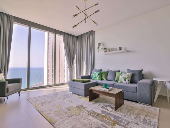 A modern living room with a grey sofa, a coffee table, and a view of the sea through large windows.