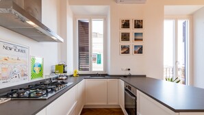 A modern kitchen with white cabinets, a black countertop, a gas stove, and a built-in oven. There are framed pictures on the wall and a window with blinds.