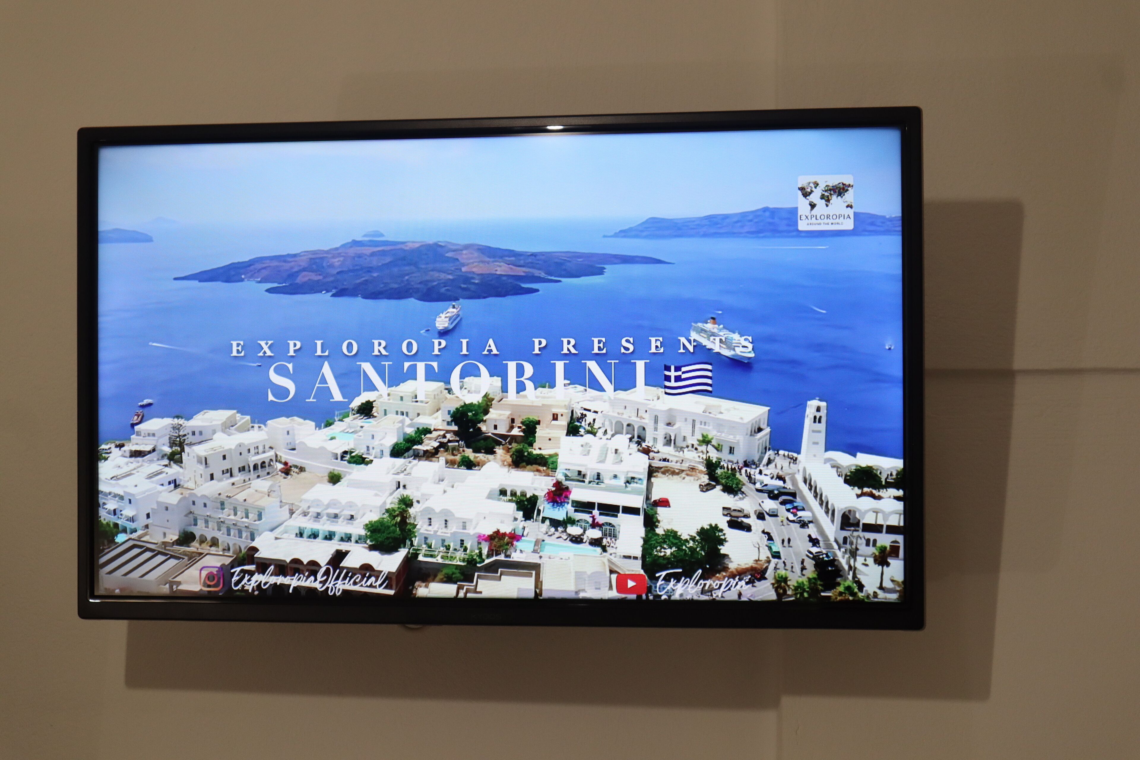 A television screen displaying a view of Santorini with the text "EXLORPIA PRESENTS SANTORINI".
