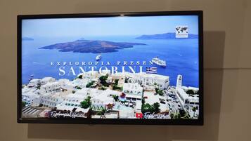 A television screen displaying a view of Santorini with the text "EXLORPIA PRESENTS SANTORINI".
