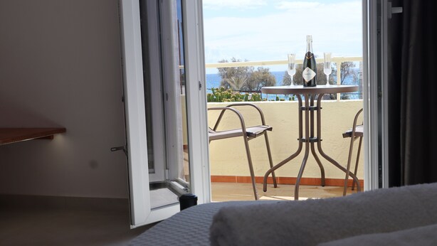 Une pièce comprenant un lit, une table avec une bouteille et des verres, et donnant sur un balcon avec des chaises et une table.