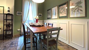 Een eetgedeelte met een houten tafel, rieten stoelen, een bloevas en ingekaderde botanische prints aan de muur.
