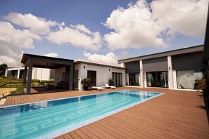 A modern outdoor pool area with a wooden deck, a clear blue pool, and a covered seating area with lounge chairs. - The Pine Mansion (Sen Monorom)