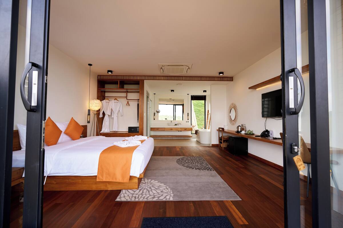 A modern hotel room with a large bed, wooden flooring, a sitting area with a view, and a TV on the wall.