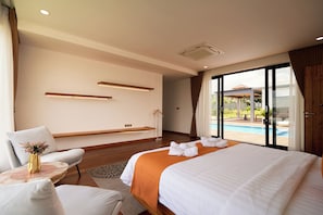 A modern hotel room with a large bed, a seating area with a chair, a small table with a vase, and a view of an outdoor swimming pool. - The Pine Mansion (Sen Monorom)