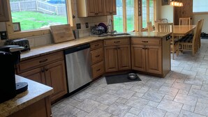 A kitchen with wooden cabinets, a dishwasher, a wooden dining table with chairs, and a large window overlooking a backyard.