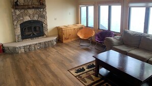 A cozy living room with a stone fireplace, wooden furniture, and large windows.