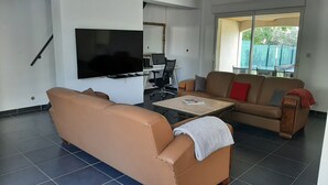A living room with a sofa, a coffee table, a TV, a laptop on a desk, and a sliding glass door leading to an outdoor area. - Location Villa Avec 2 Appartements Ind, Proche du Lac de Ste Croix et du Verdon (Régusse)