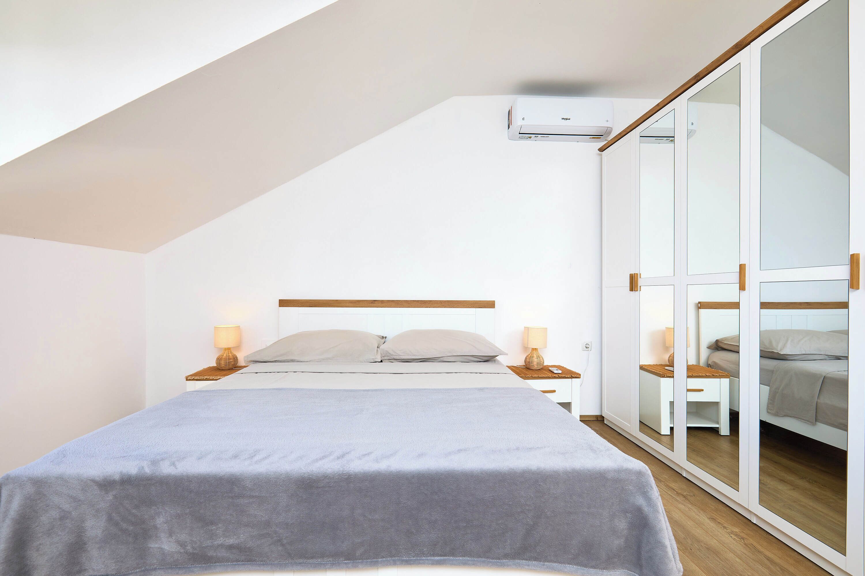 A modern bedroom with a sloped ceiling, a bed with a grey bedspread, bedside lamps, and a large mirror cabinet.