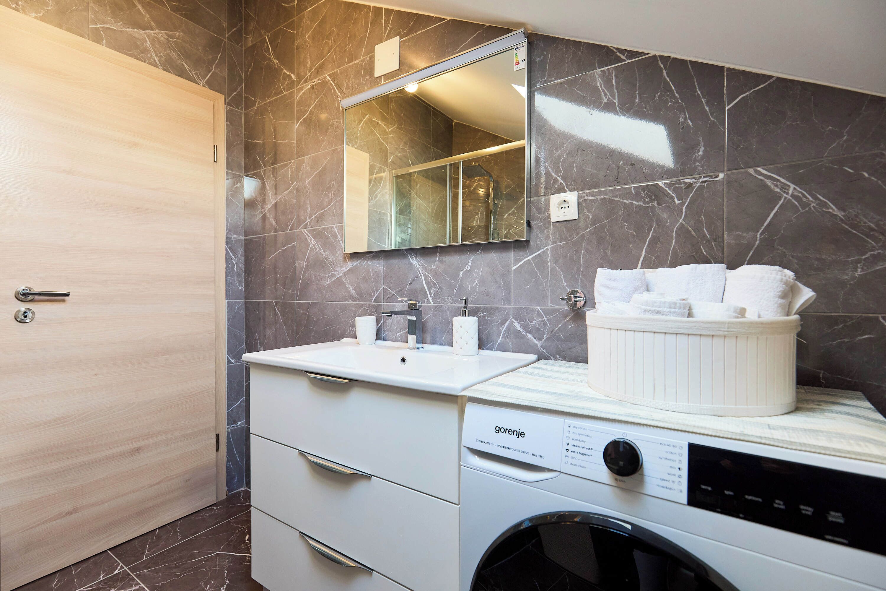 Ein modernes Badezimmer mit Marmorverkleidung, einem Waschbecken mit Spiegel darüber, einer Waschmaschine und einer Holztür.