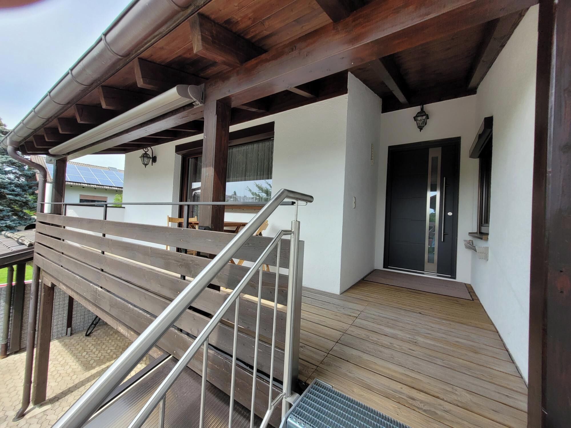 Une terrasse en bois avec une rambarde en métal, un espace couvert avec une porte, et une vue sur l’extérieur.