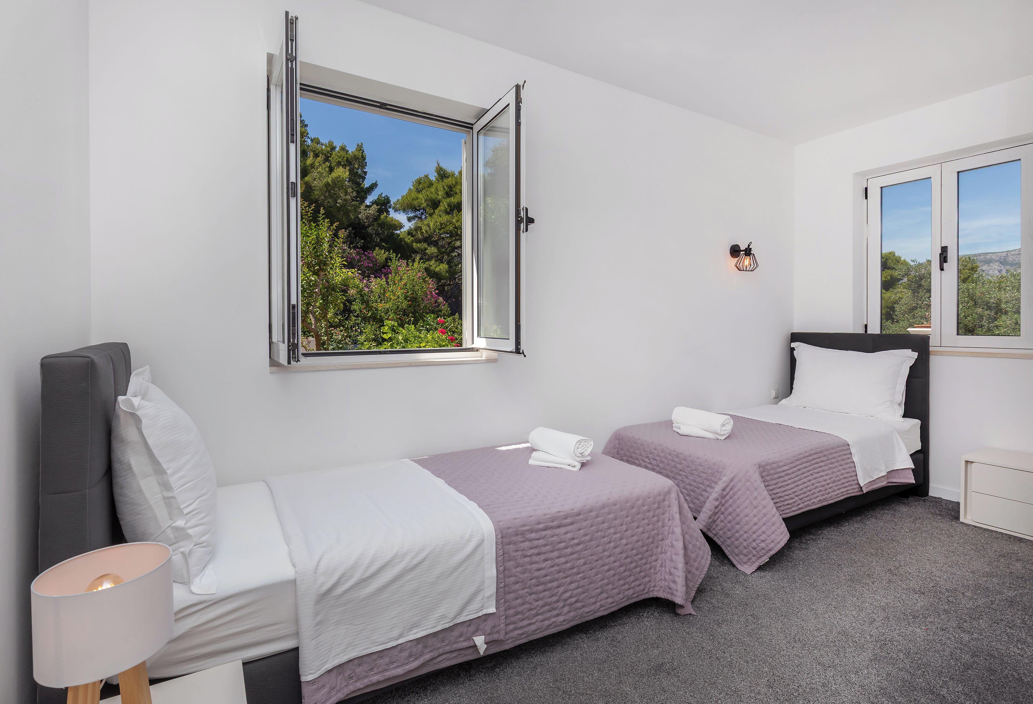 Ein Schlafzimmer mit zwei Betten, einem Fenster, einer Lampe und Blick auf Bäume.