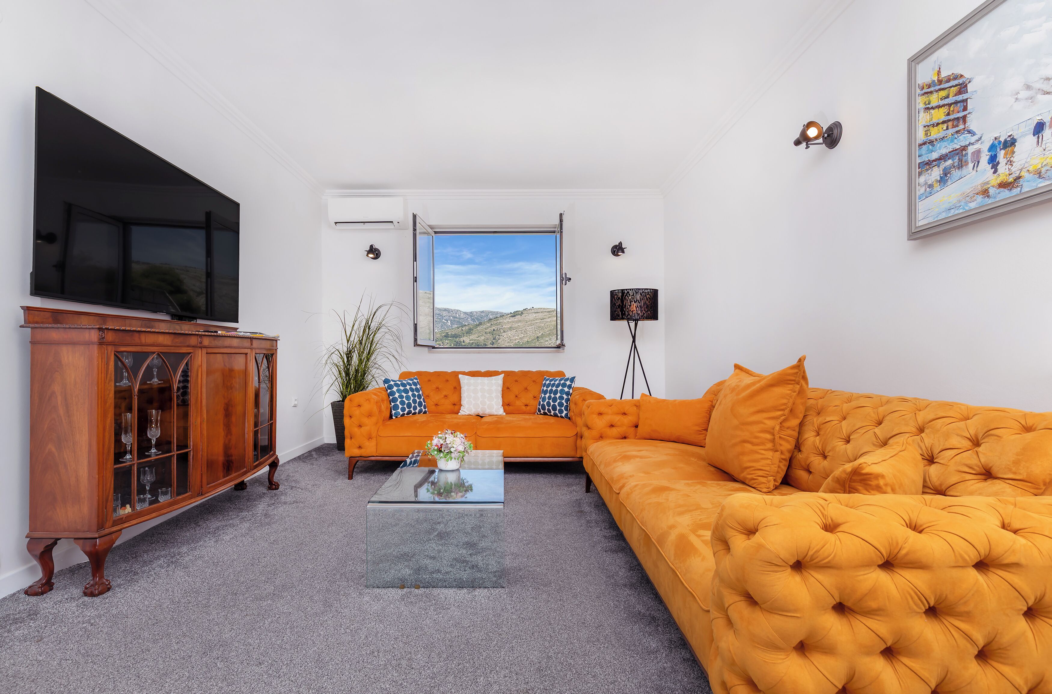 A living room with a large flat-screen TV, a sofa, a coffee table, and a window with a view of hills.